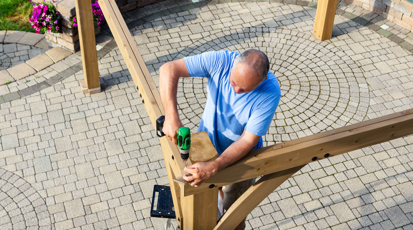 Technicien professionnel installant une pergola bioclimatique en aluminium sur terrasse résidentielle, visseuse en main, structure partiellement montée avec lames orientables